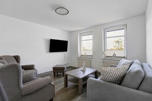 Una sala moderna con un televisor de pantalla plana montado en la pared, una mesa de centro de madera, sofás grises y un pequeño otoman.