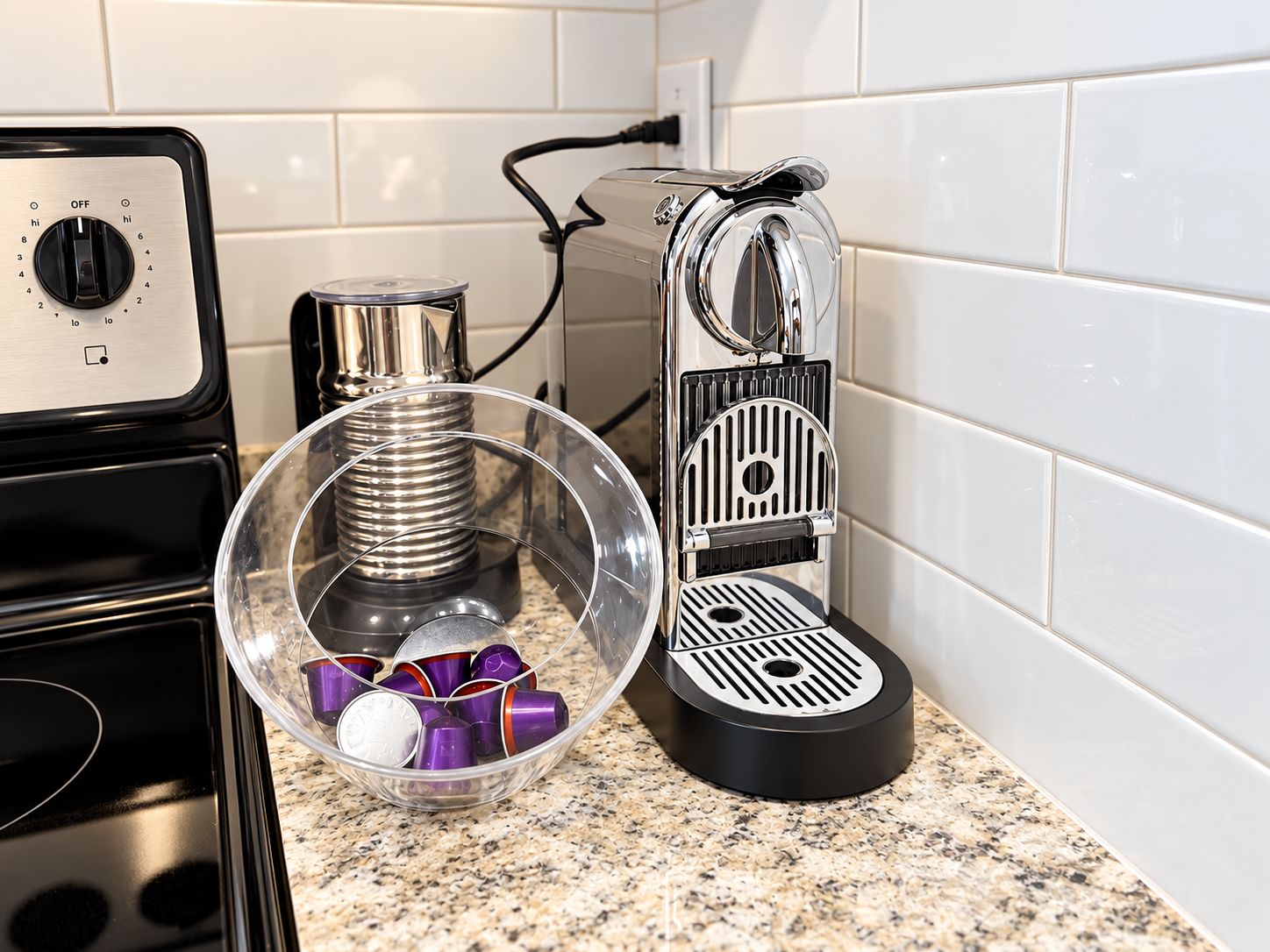 Una cocina con cafetera, hervidor de agua y un recipiente de vidrio con cápsulas de café sobre una encimera de granito.