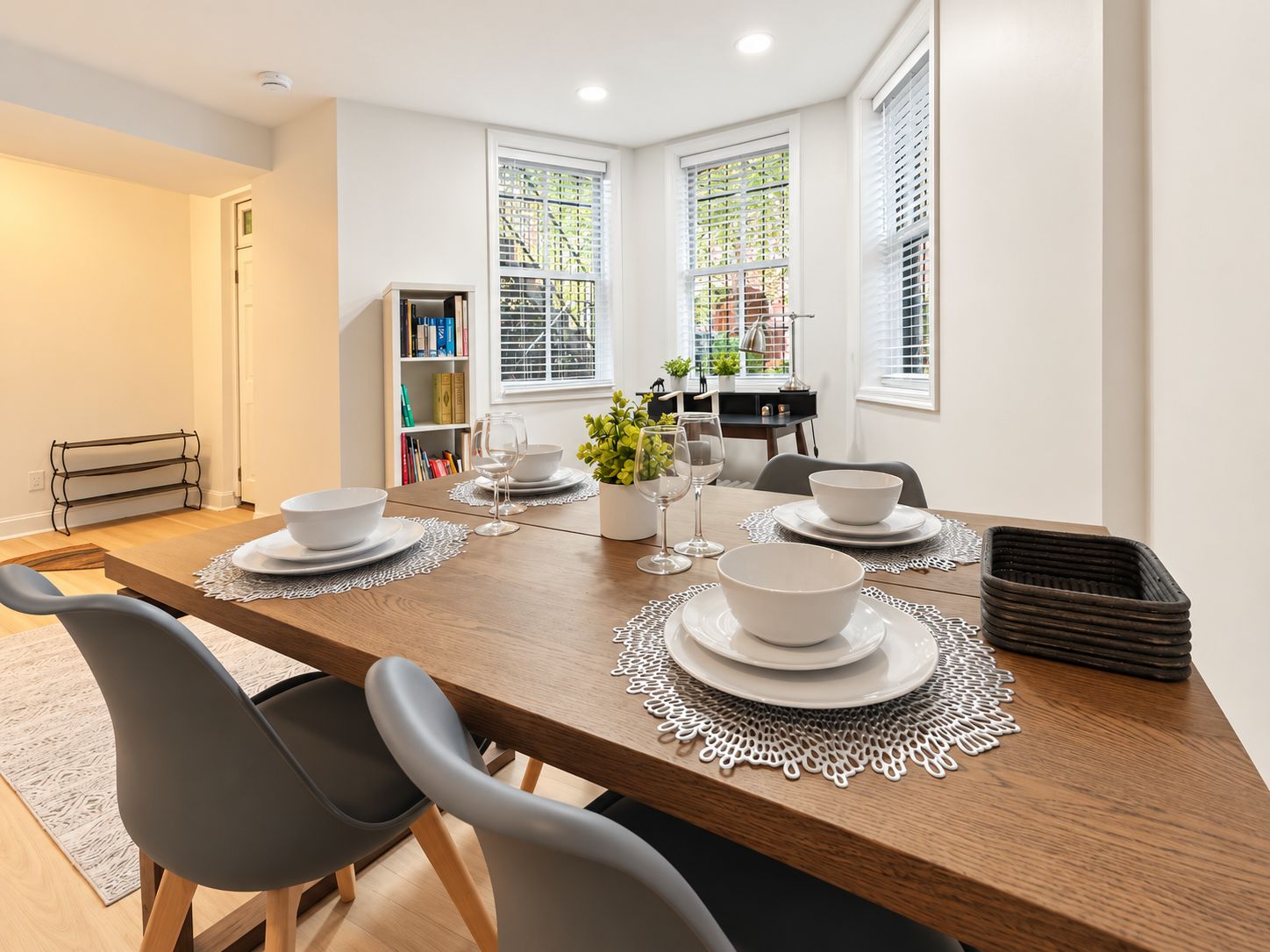 A dining area with a wooden table set for four, surrounded by chairs, with a bookshelf and a window with blinds in the background.