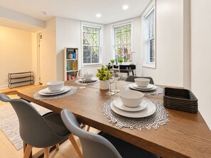 A dining area with a wooden table set for four, surrounded by chairs, with a bookshelf and a window with blinds in the background. - 1BR in Logan Circle - DC Metro - We Host (Washington)