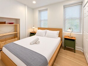 A neatly made bed with white linens and a folded towel, a bedside table with a lamp, and a shelf with a red bowl. - 1BR in Logan Circle - DC Metro - We Host (Washington)