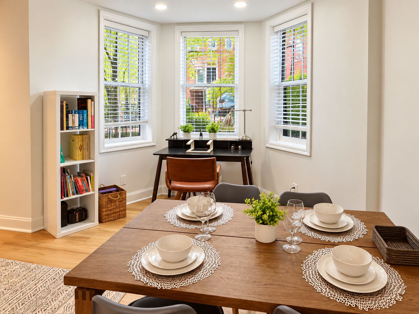 Comedor con mesa de madera dispuesta para cuatro personas, rodeado de paredes blancas y ventanas amplias con persianas.