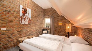 Un dormitorio con pared de ladrillo, cama con ropa de cama blanca, una silla de mimbre, una lámpara de pie y una ventana con cortinas.