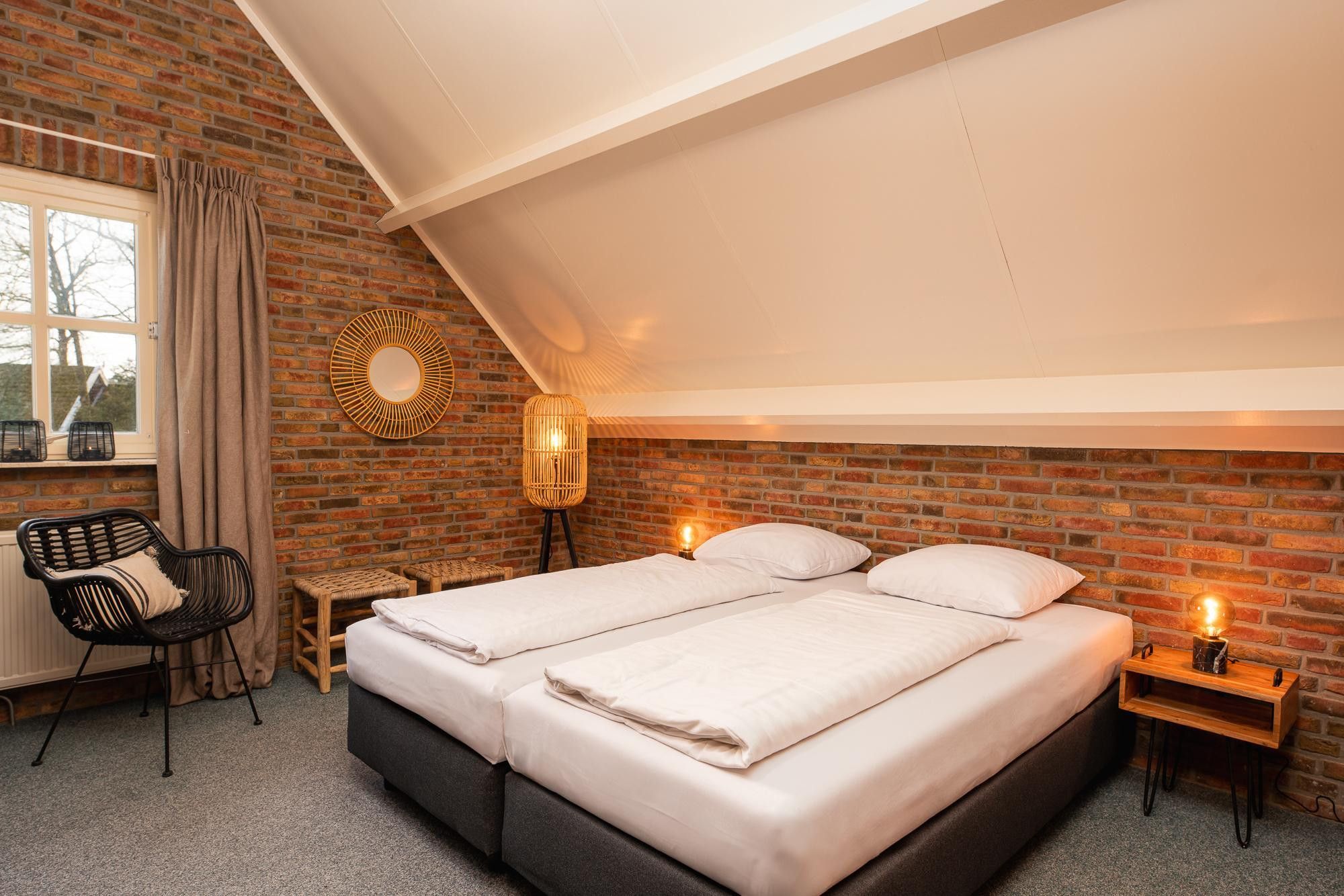 A bedroom with a brick wall, a large bed, a chair, a bedside table, and a window with curtains.