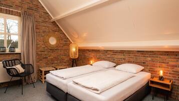 A bedroom with a brick wall, a large bed, a chair, a bedside table, and a window with curtains.