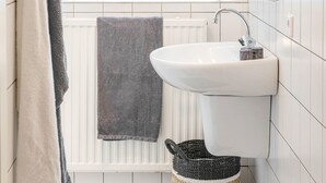 Un baño con un lavamanos blanco, un perchero con dos toallas, un radiador y una pequeña estantería con una planta.