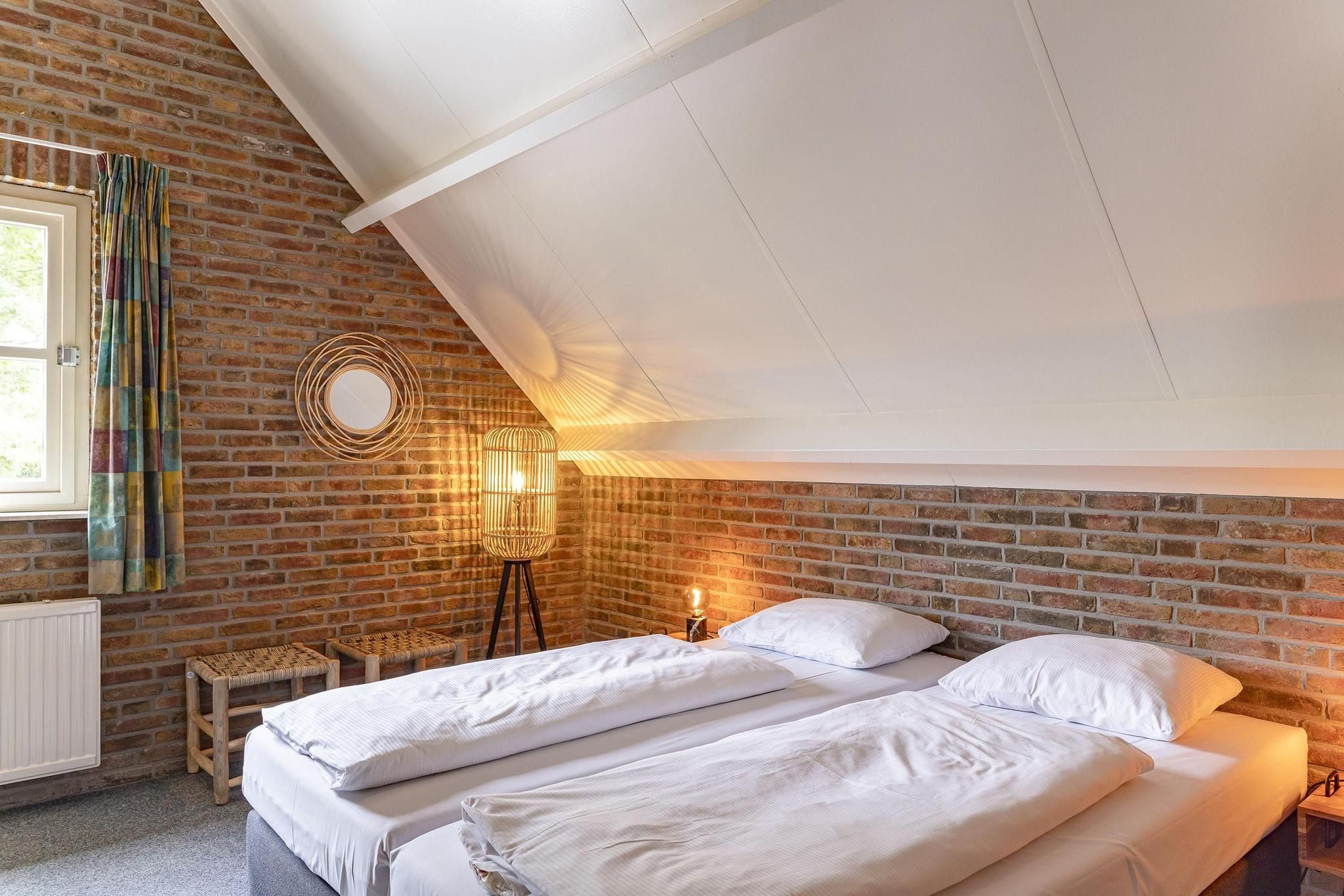 Un dormitorio con techo inclinado, pared de ladrillo, dos camas, una ventana con cortinas y una lámpara de pie.