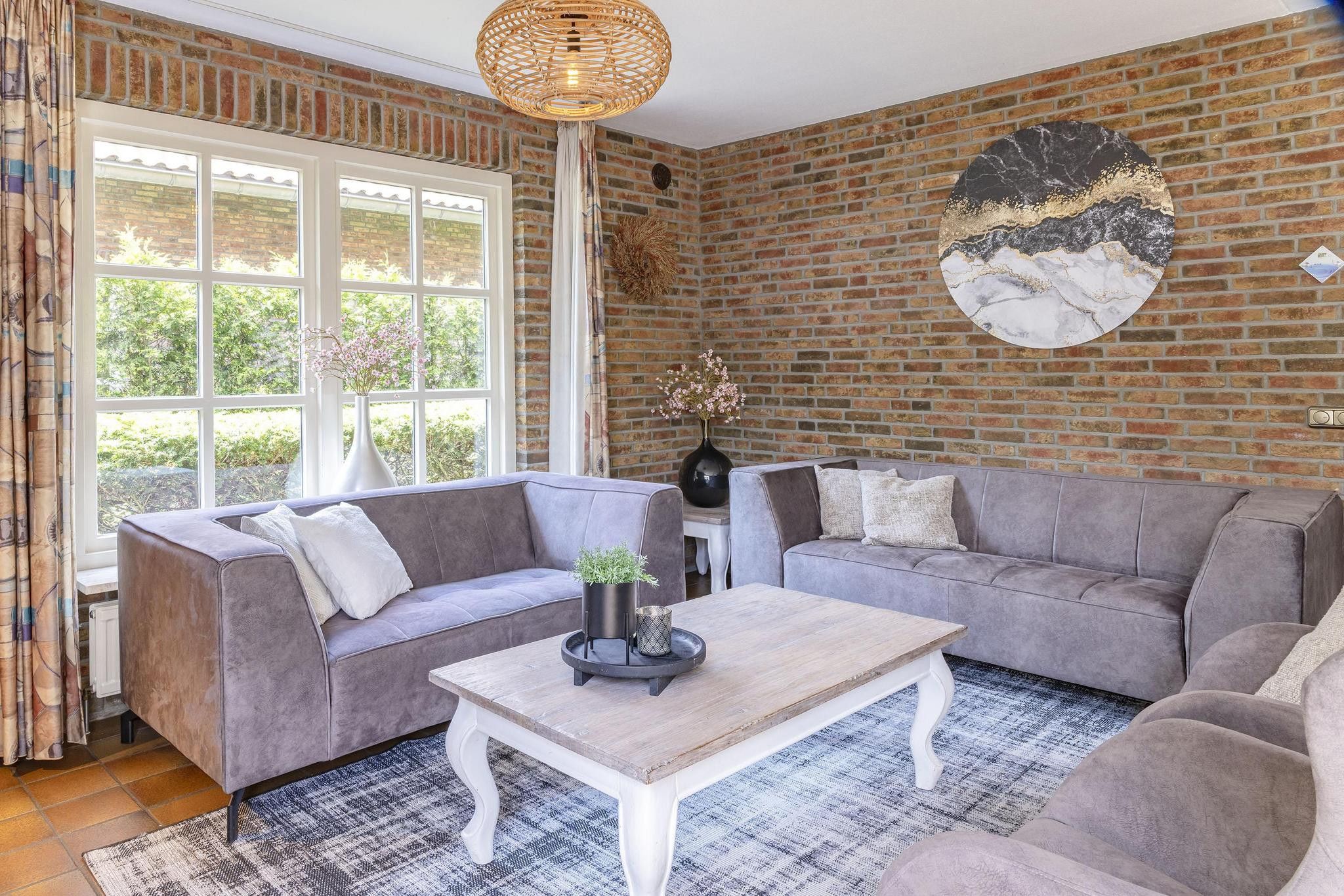 Een woonkamer met een bakstenen muur, twee grijze banken, een witte salontafel en een hangende lamp.