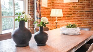 Una mesa de madera con dos jarrones oscuros que contienen flores blancas, una lámpara y una pila de platos blancos sobre un fondo de pared de ladrillo.