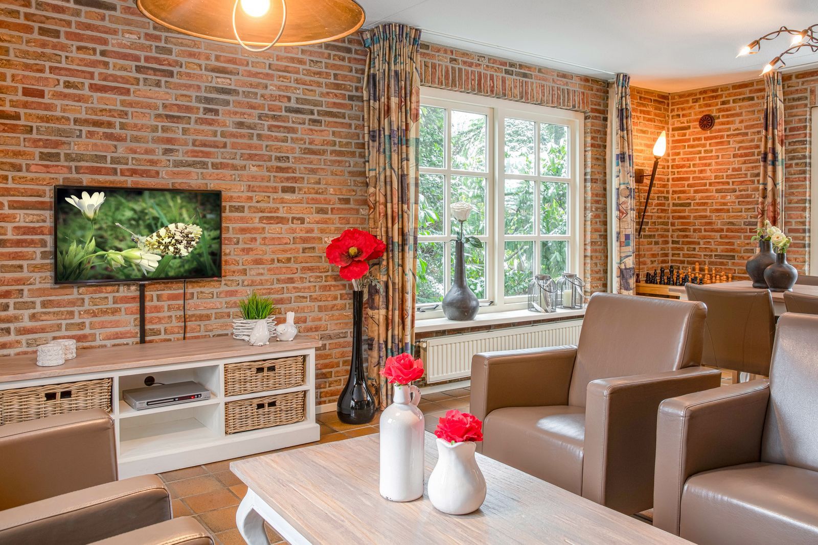 A living room with a brick wall, a television, a window with curtains, a chessboard, and a table with vases.