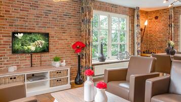 A living room with a brick wall, a television, a window with curtains, a chessboard, and a table with vases.