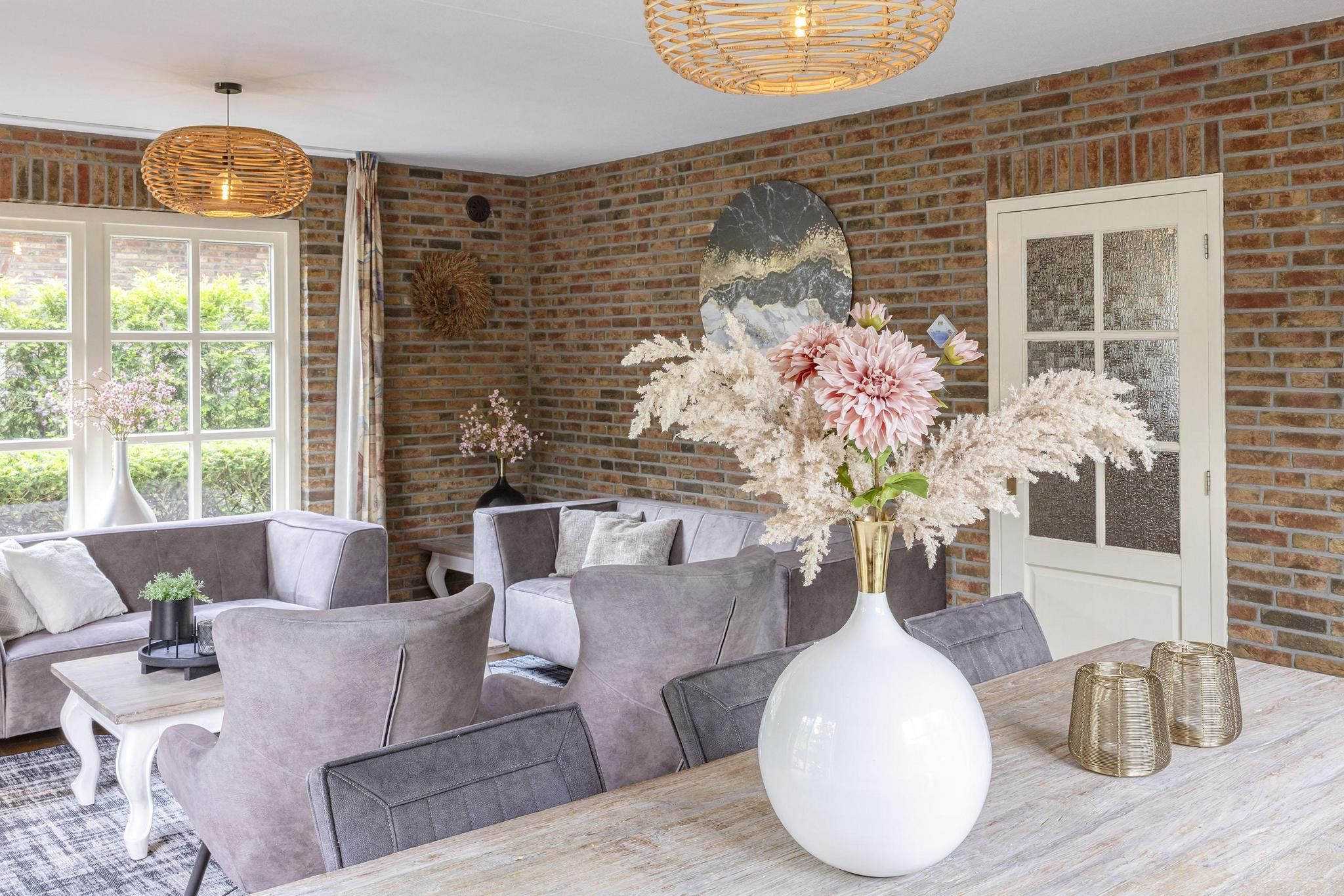 Un coin repas avec un mur en briques, un grand vase blanc orné de fleurs et des meubles recouverts d’un tissu gris.