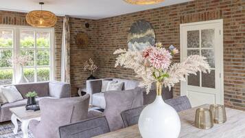 Un coin repas avec un mur en briques, un grand vase blanc orné de fleurs et des meubles recouverts d’un tissu gris.