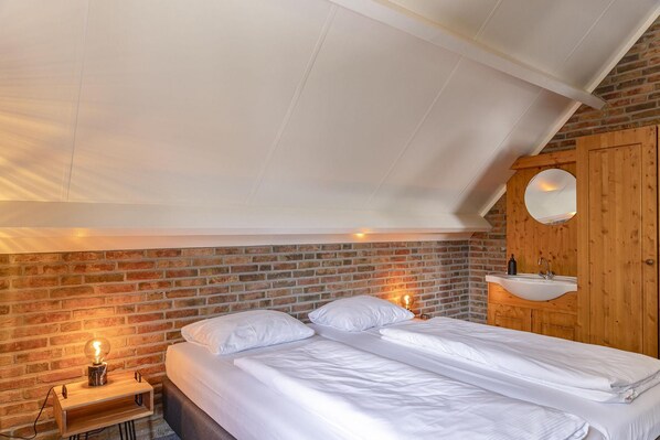 A bedroom with a brick wall, a wooden door, a round mirror, a white bed, and a bedside table with a lamp.