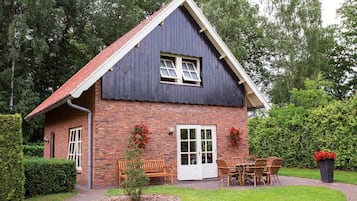 Een tweelaags huis met een bakstenen gevel, een rode dakbedekking en een kleine voortuin met een bankje en een tafel.