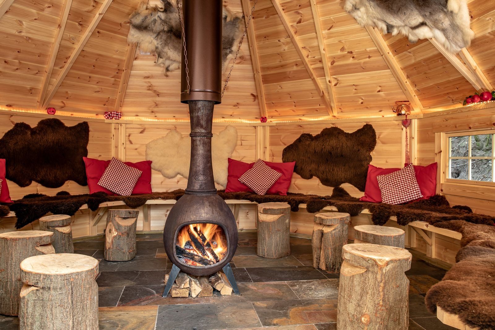 A log fire stove in a wooden cabin with fur rugs and pillows.