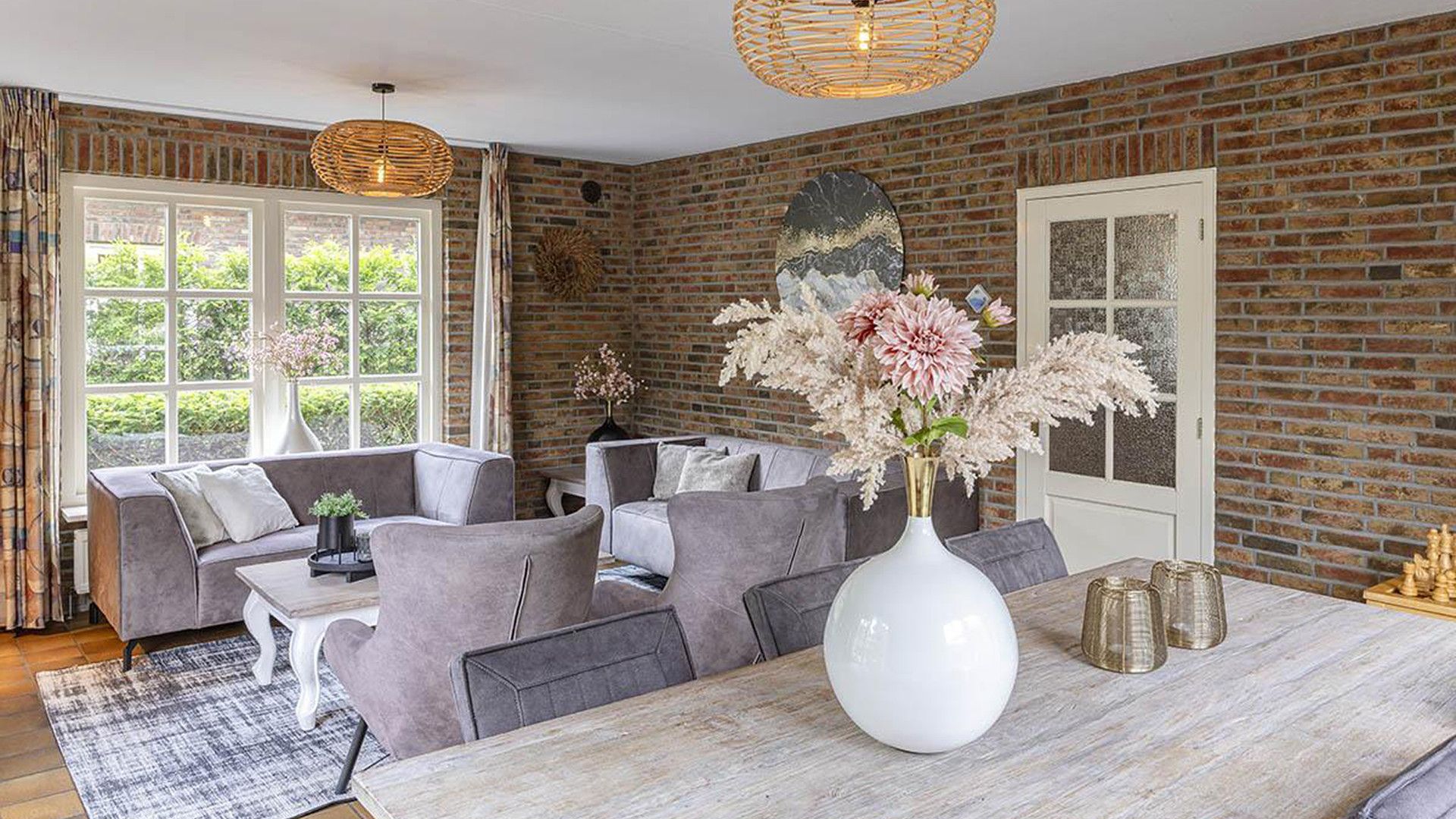 Een moderne woonkamer met een bakstenen muur, grijze banken, een eettafel en een groot raam met gordijnen.