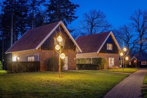 Une maison à deux étages, avec une façade en briques et un toit recouvert de tuiles rouges, entourée d’une pelouse bien entretenue et d’arbres.