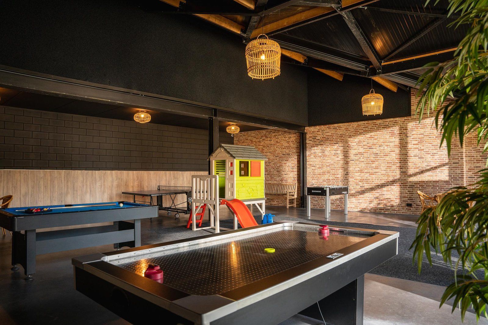 Una mesa de billar con un disco de neón, una casita de juegos para niños y una mesa de ping pong en una habitación con paredes de ladrillo y vigas de madera.