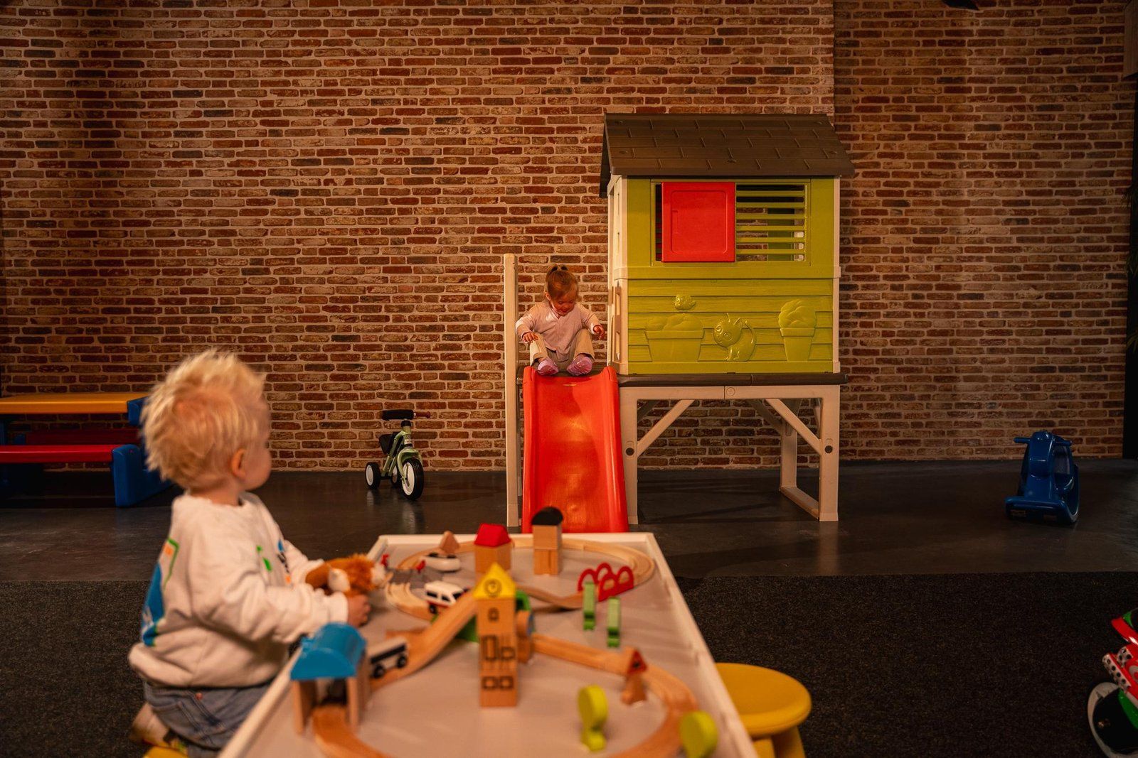 A child playing with toys in a room with a brick wall and a playhouse.