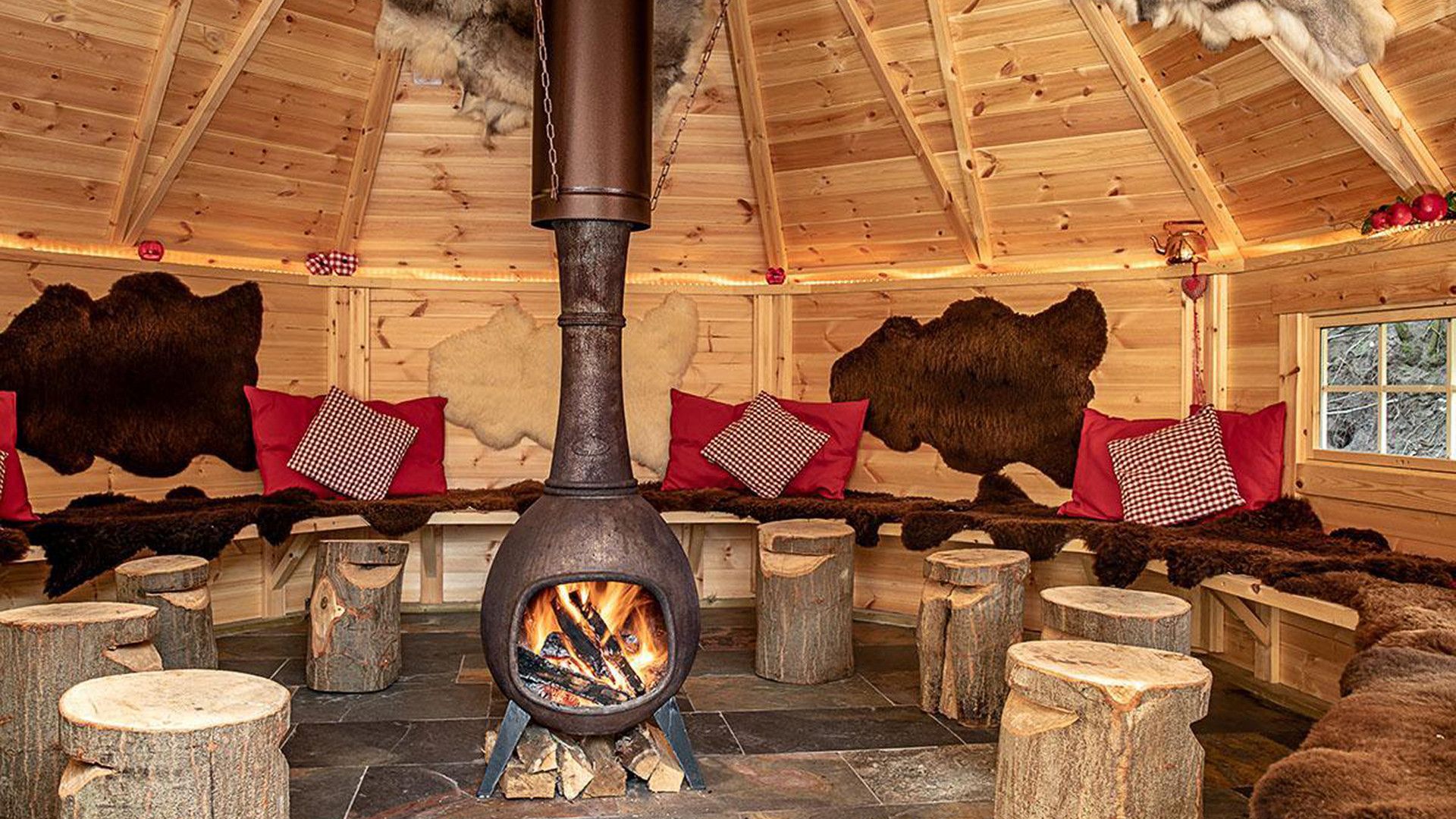 Interior rústico de cabaña con estufa de leña en el centro, paredes de madera y pieles de oso como decoración.