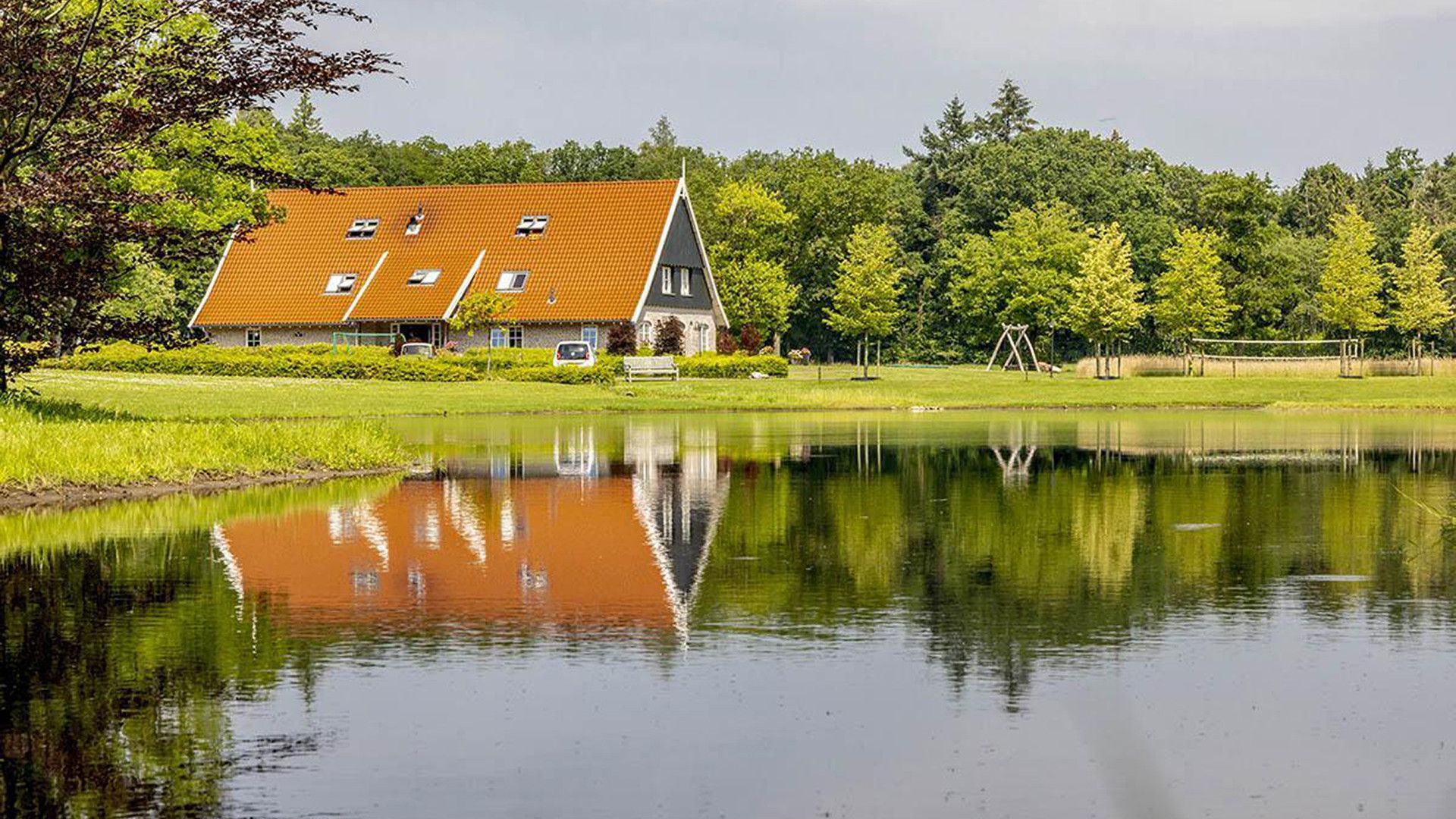 Une maison avec un toit rouge, un étang devant et des arbres en arrière-plan.