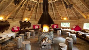 A cozy log cabin interior with a central stone fireplace, wooden benches, and fur decorations.