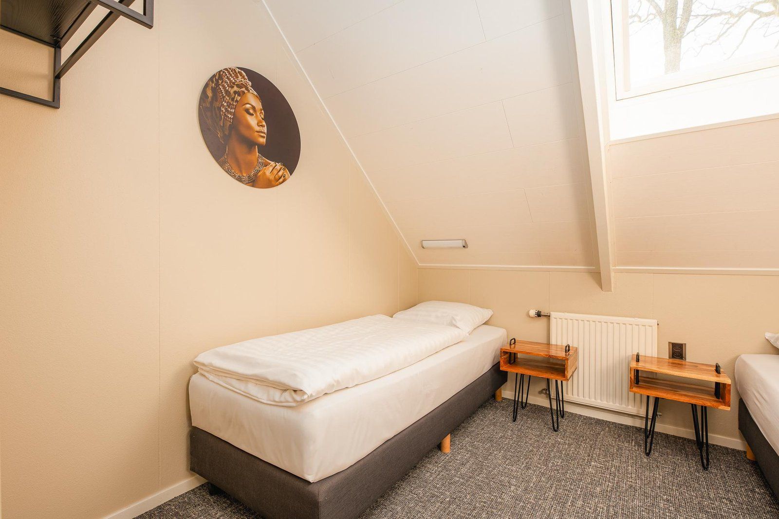Une petite chambre avec un lit simple, un plafond en pente, un radiateur, une table de chevet et un tableau accroché au mur.