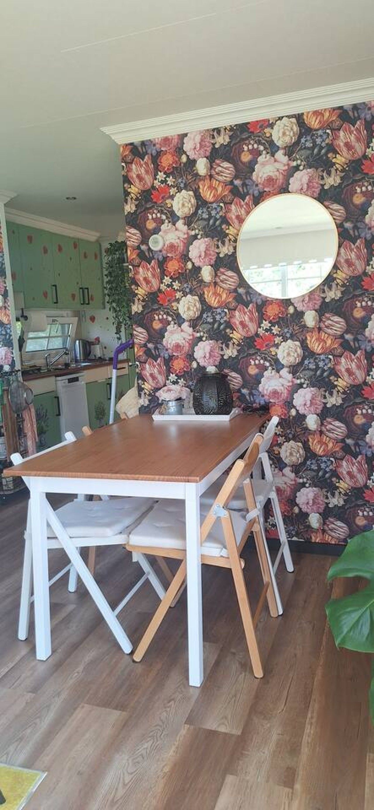 Een eetgedeelte met een houten tafel en stoelen, bloemenbehang en een ronde spiegel aan de muur.