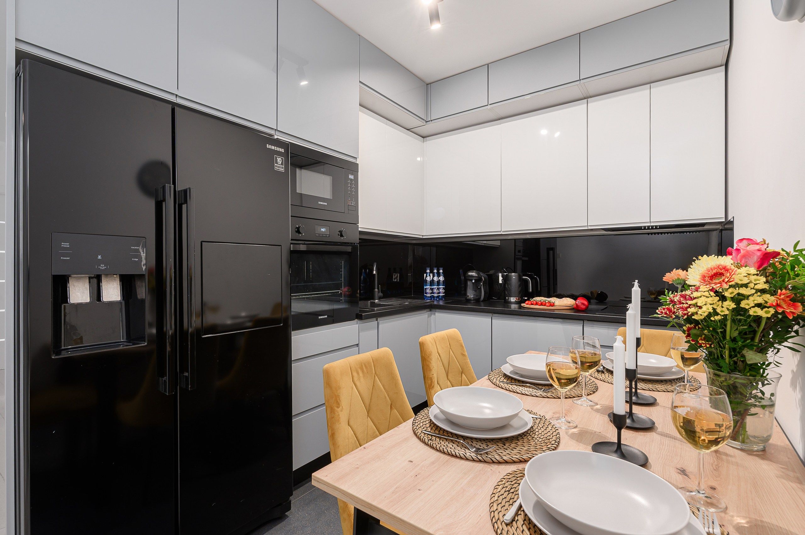 A modern kitchen with a black refrigerator, white cabinets, and a dining table set with plates, glasses, and a vase of flowers.