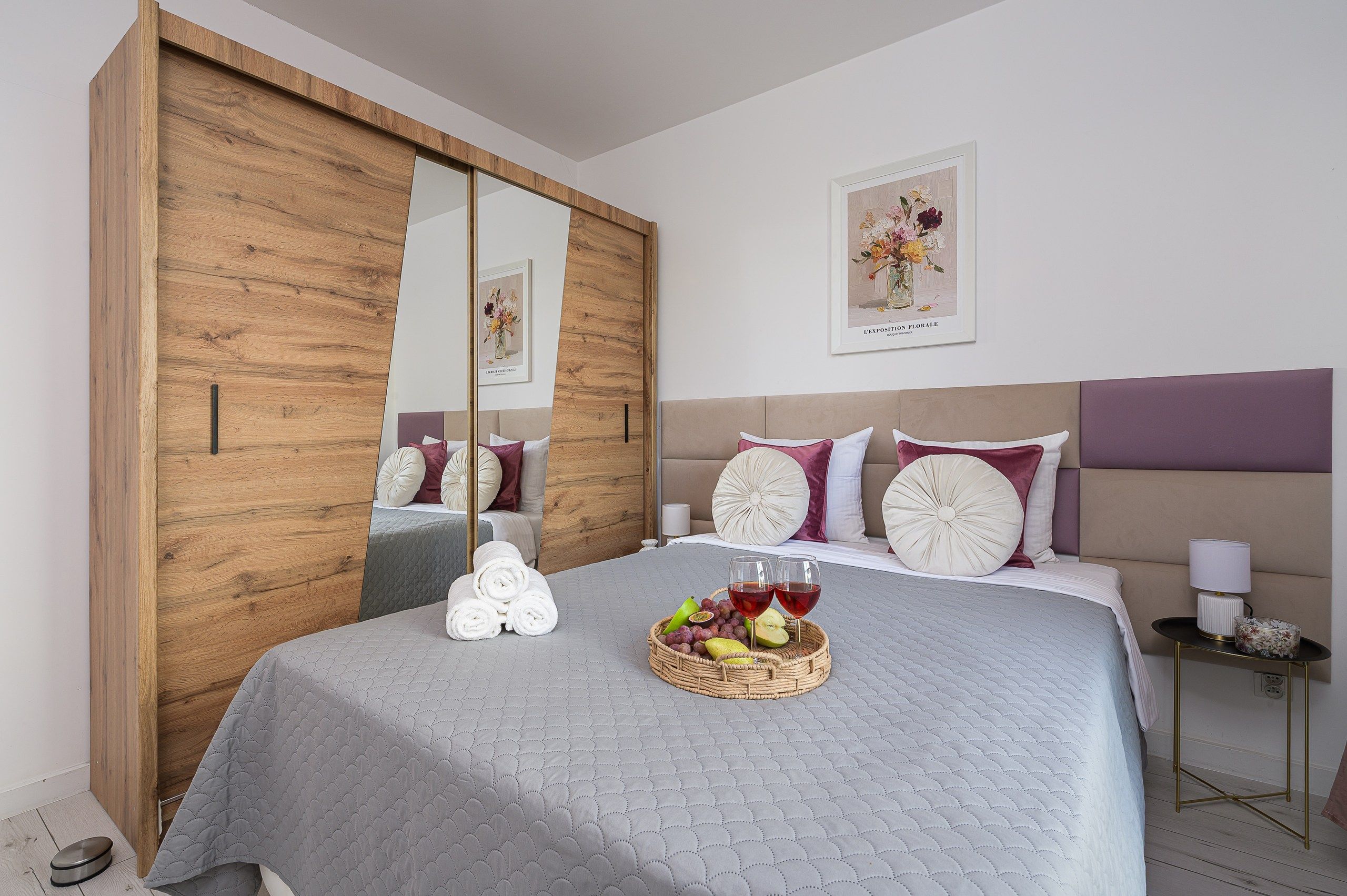 A bedroom with a large bed, a wooden wardrobe, and a mirror. There is a framed picture on the wall and a small table with a lamp.