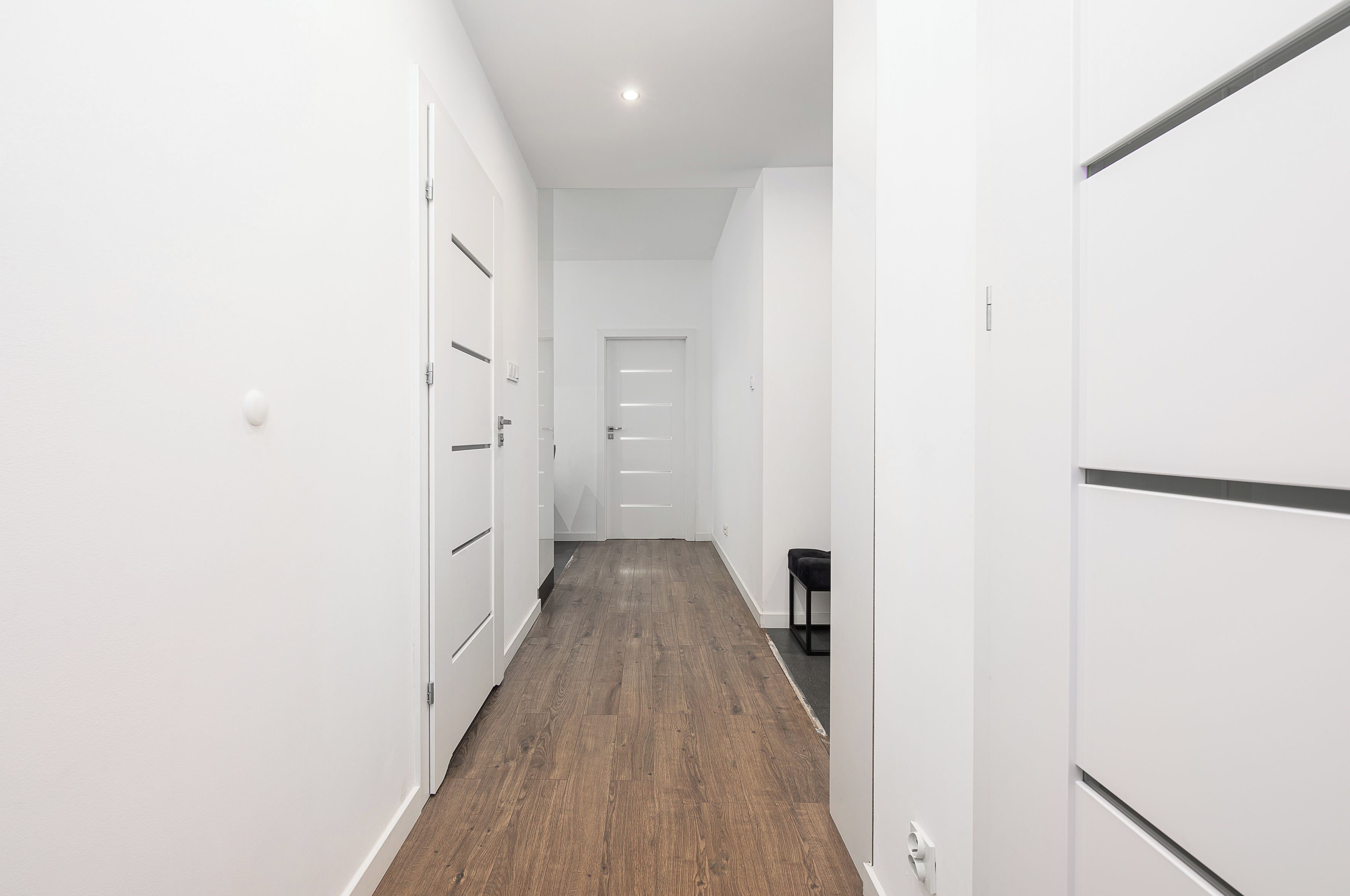 A narrow hallway with white walls and wooden flooring, leading to a door at the end.
