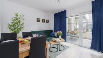 A modern living room with a dining area, a green sofa, a dining table with chairs, a potted plant, and a view through a glass door.