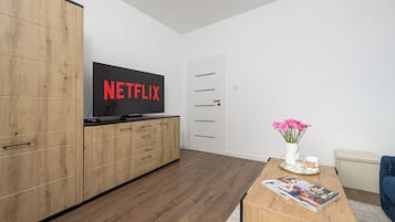 Una sala de estar moderna con un mueble de madera, un televisor de pantalla plana mostrando 'NETFLIX', una puerta blanca, una mesa de centro con revistas y un jarrón con flores rosas.