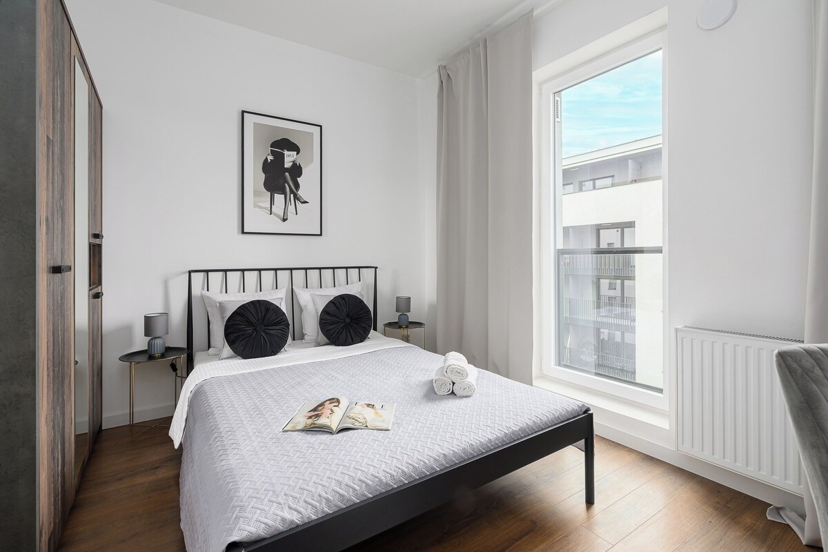 A modern bedroom with a bed, a window, a framed picture, and a bedside table.