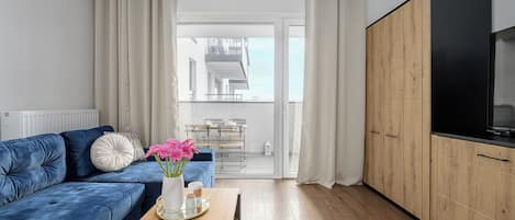 A modern living room with a blue sofa, a coffee table with magazines, and a view to a balcony with a dining area.