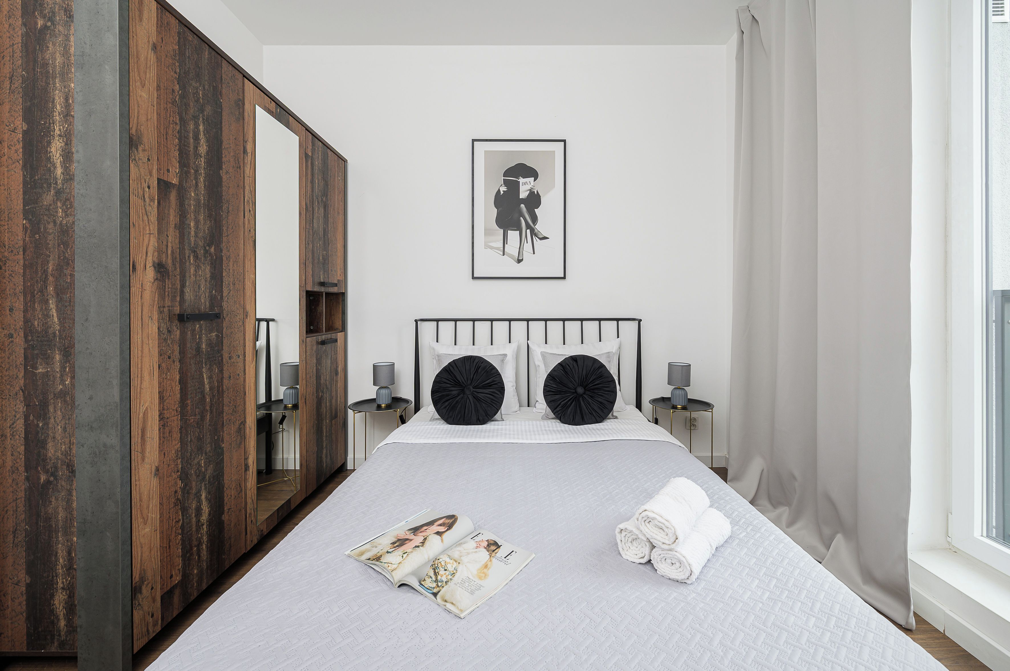 Un dormitorio ordenado con un armario grande, un cuadro en la pared, una cama con dos almohadas negras, una revista y toallas enrolladas.
