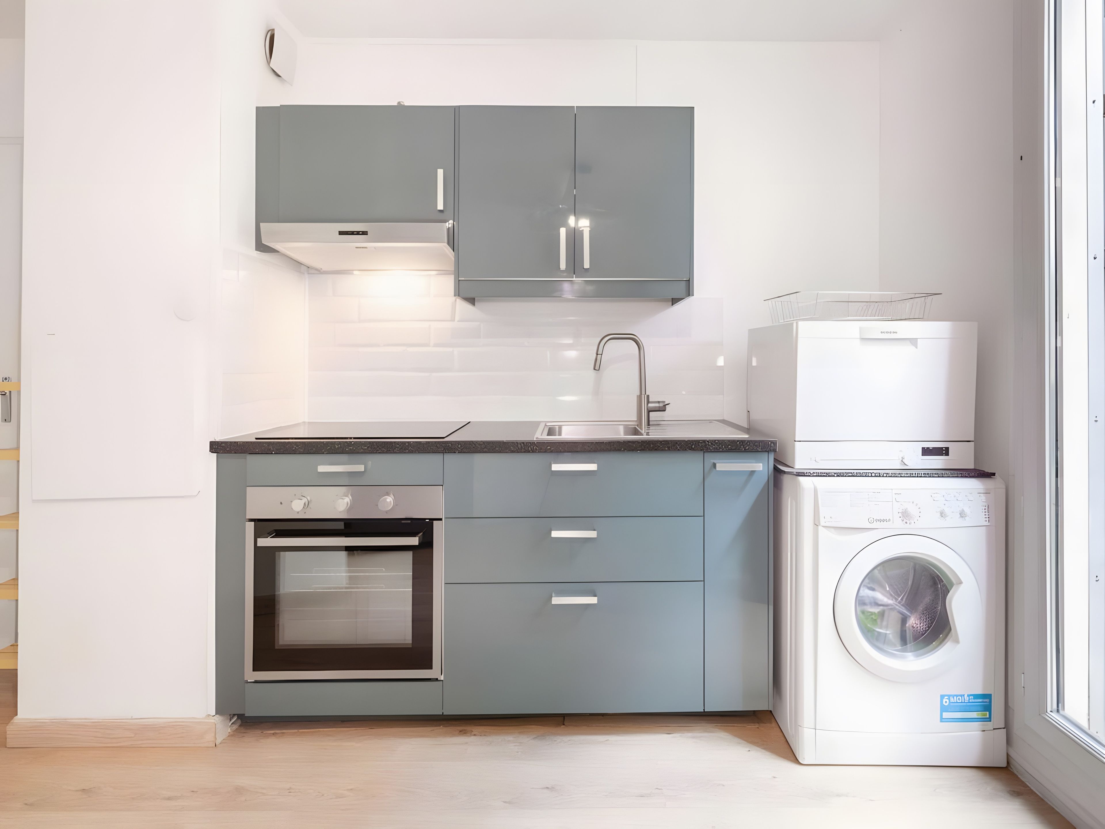 Een kleine keuken met grijze kastjes, een inbouwoven, een spoelbak en een gestapelde wasmachine en droger.