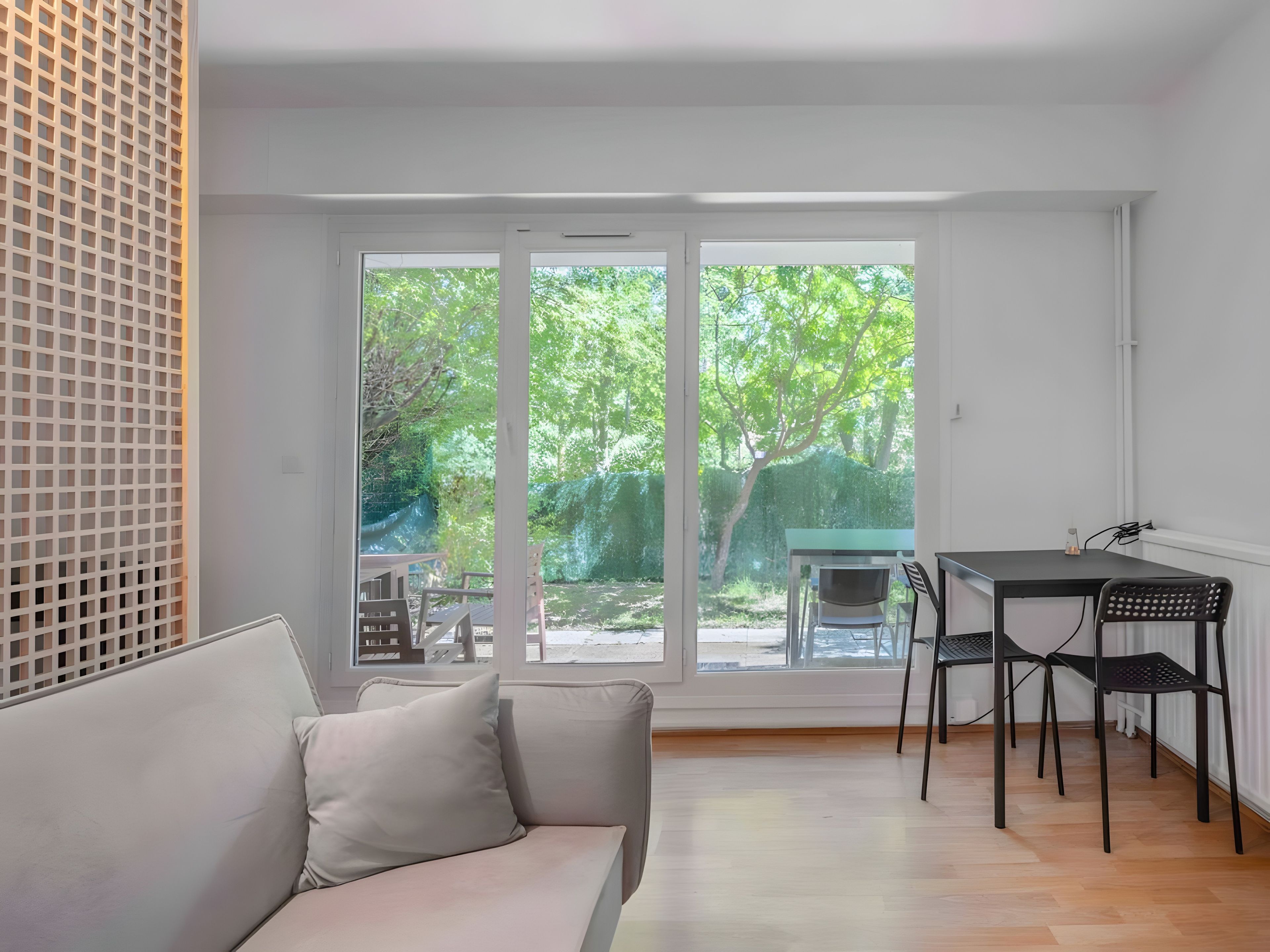 A room with a sofa, a dining table with chairs, and large windows with a view of greenery.