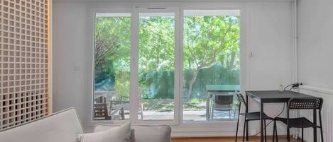 A room with a sofa, a dining table with chairs, and large windows with a view of greenery.