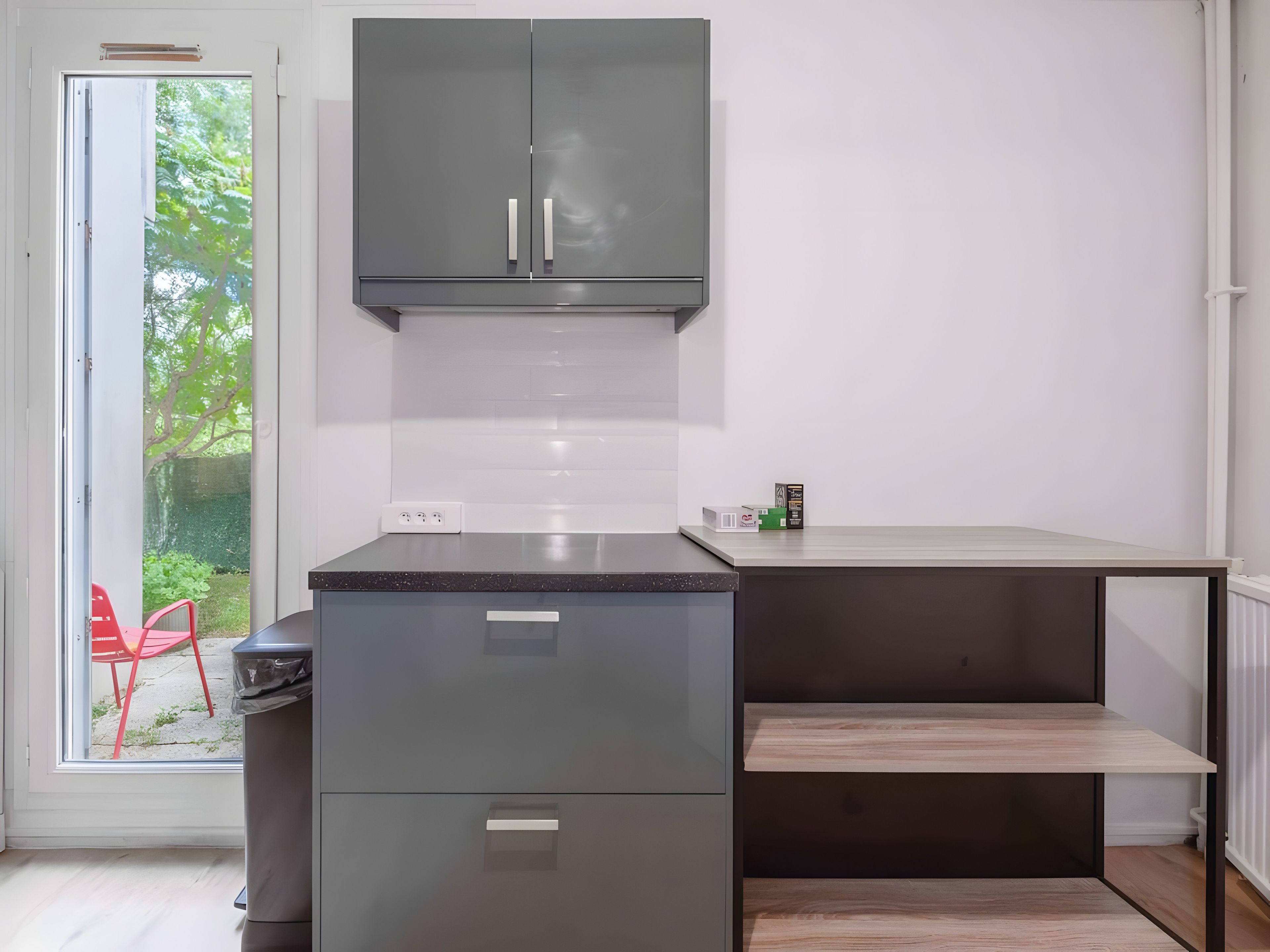 A kitchen with a grey cabinet, a countertop, and a view of a red chair outside.