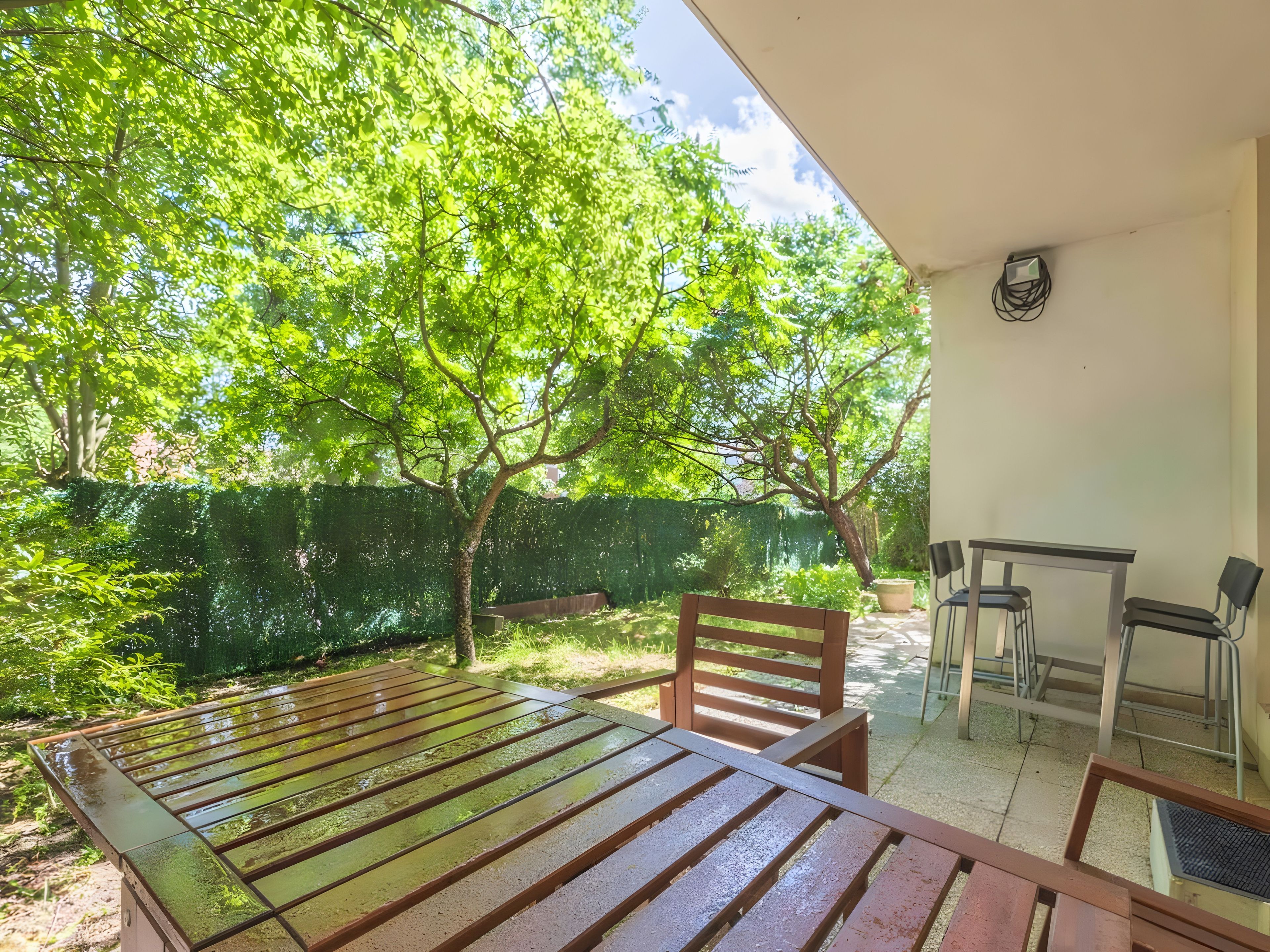 An outdoor patio with a table and chairs, surrounded by trees and a green fence.