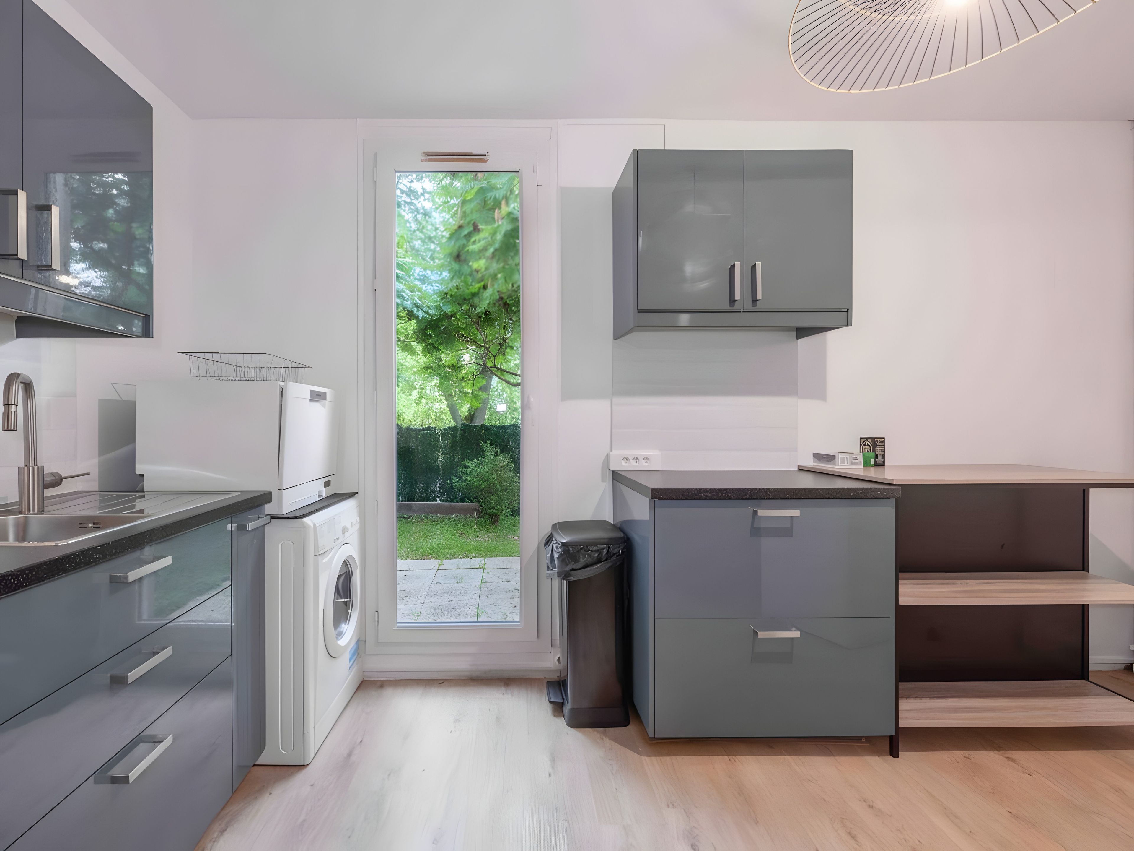 A modern kitchen with a washing machine, grey cabinets, and a view of greenery outside.