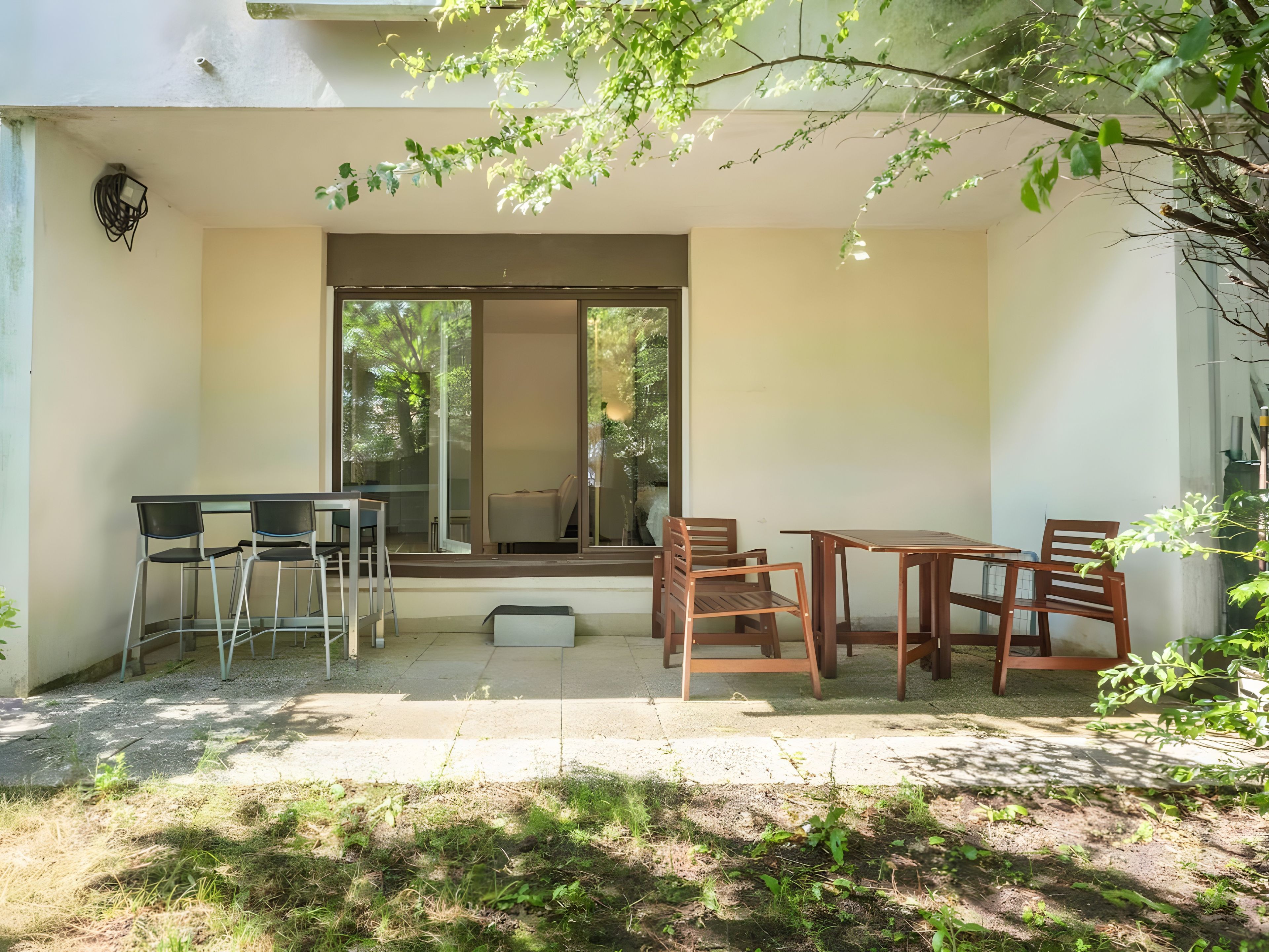 Un área de patio con mesa y sillas, una pequeña mesa con sillas, y vista al exterior a través de un ventanal de vidrio.