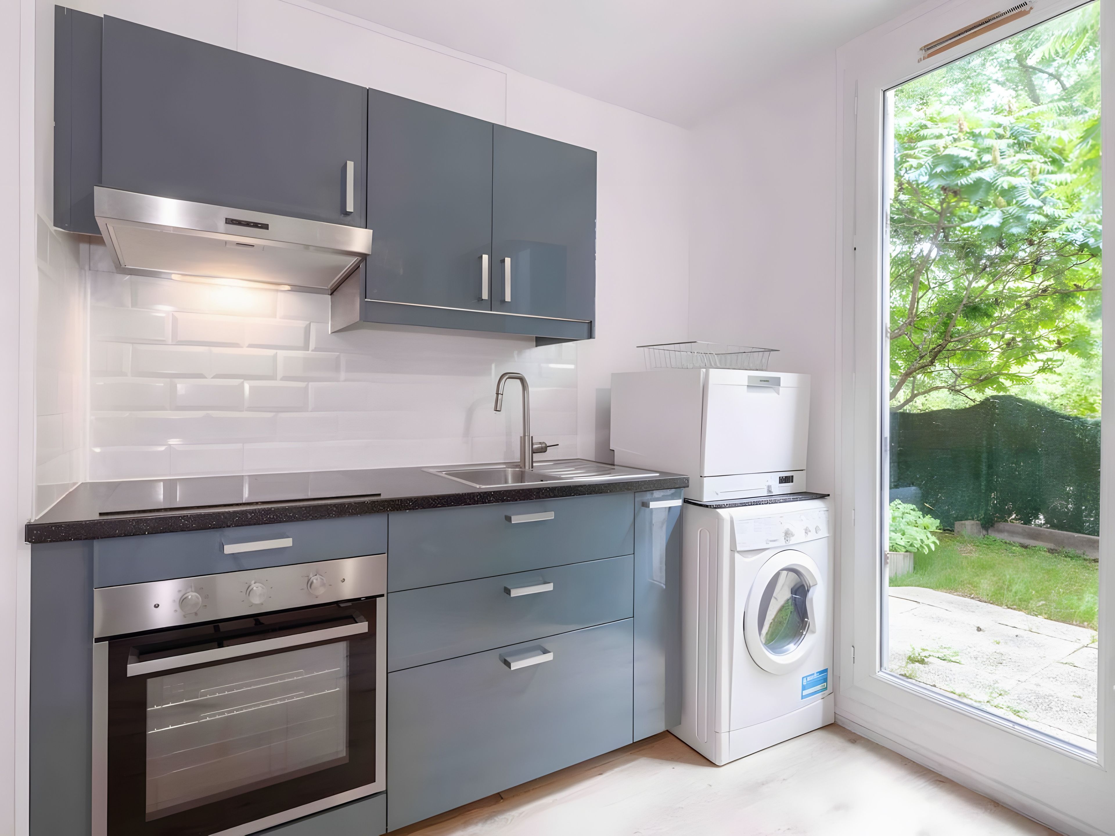 A modern kitchen with grey cabinets, a stainless steel range hood, a built-in oven, a sink, and a front-loading washing machine.
