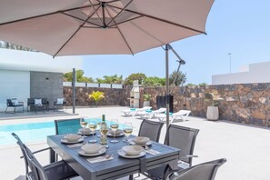 An outdoor dining area with a table set for two, a pool, and a stone wall in the background. - Villa Malvasia Sol by Villa Plus (Playa Blanca)