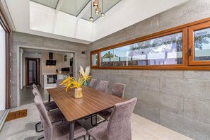 A dining area with a wooden table and cushioned chairs, a vase with yellow flowers, and a view of the outside through a large window. - Villa Malvasia Sol by Villa Plus (Playa Blanca)