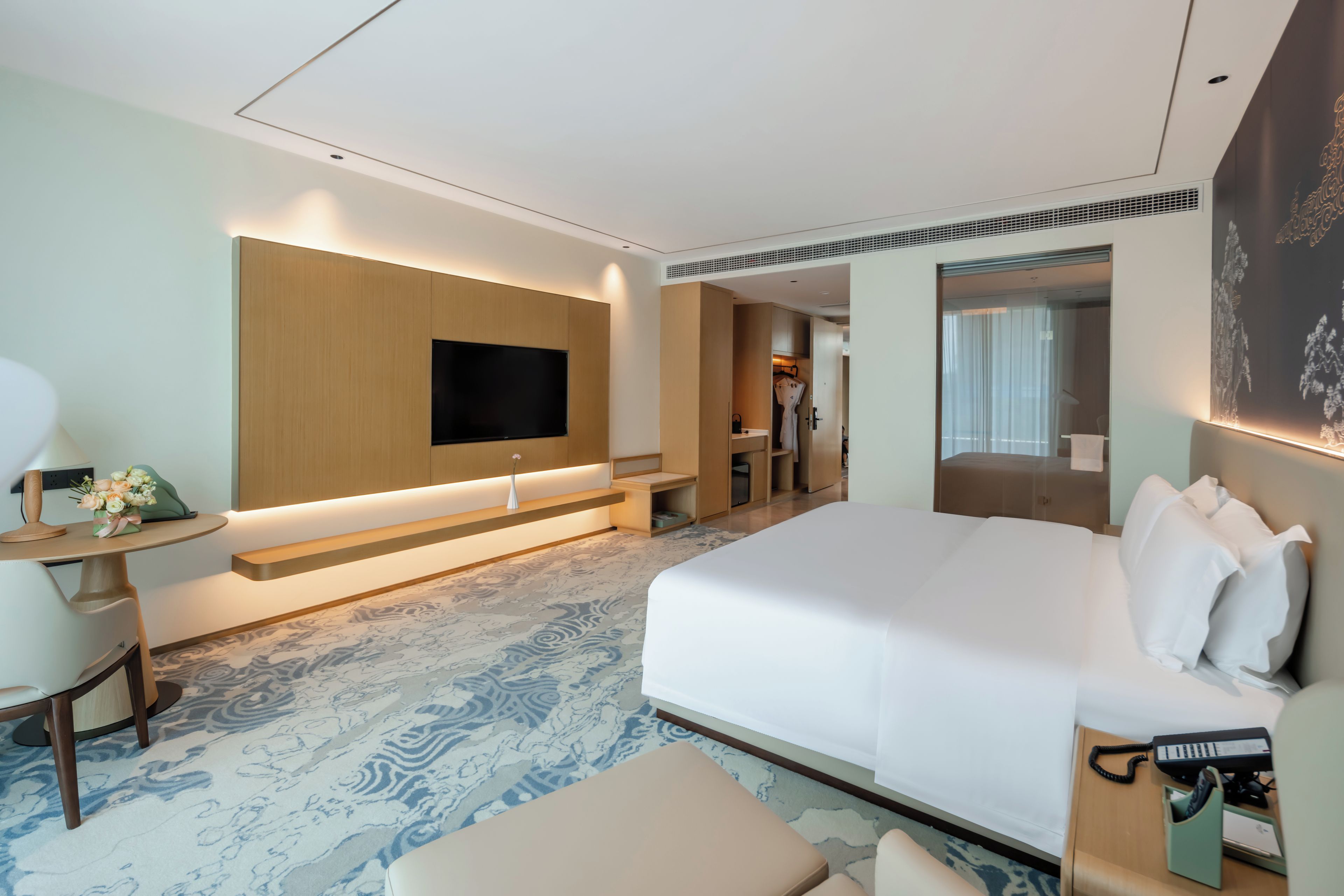 Habitación de hotel moderna con una cama grande, un televisor de pantalla plana en la pared, un escritorio con lámpara, y un baño visible a través de una puerta abierta.