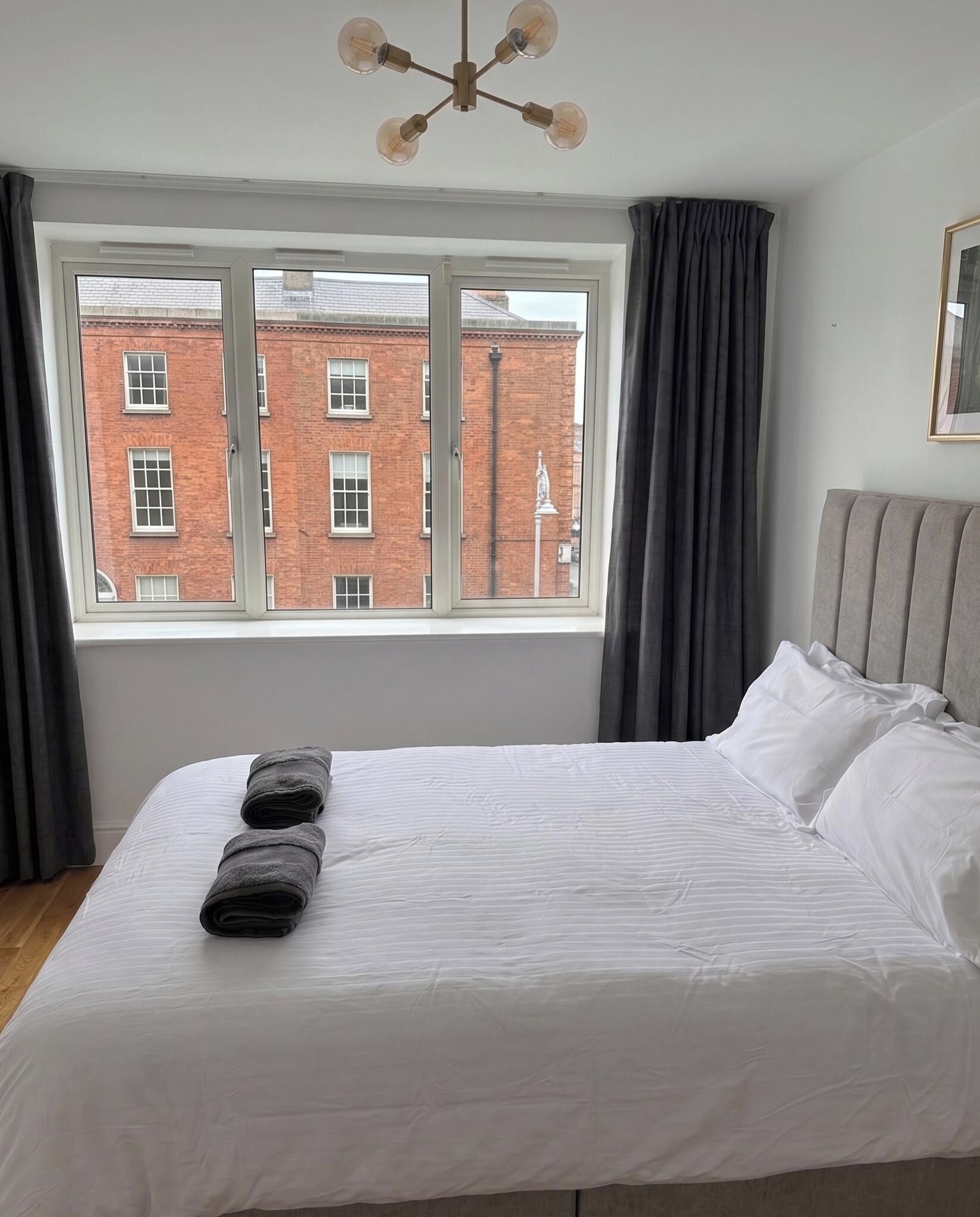 A bedroom with a large window, a bed with white linens, and a grey headboard.