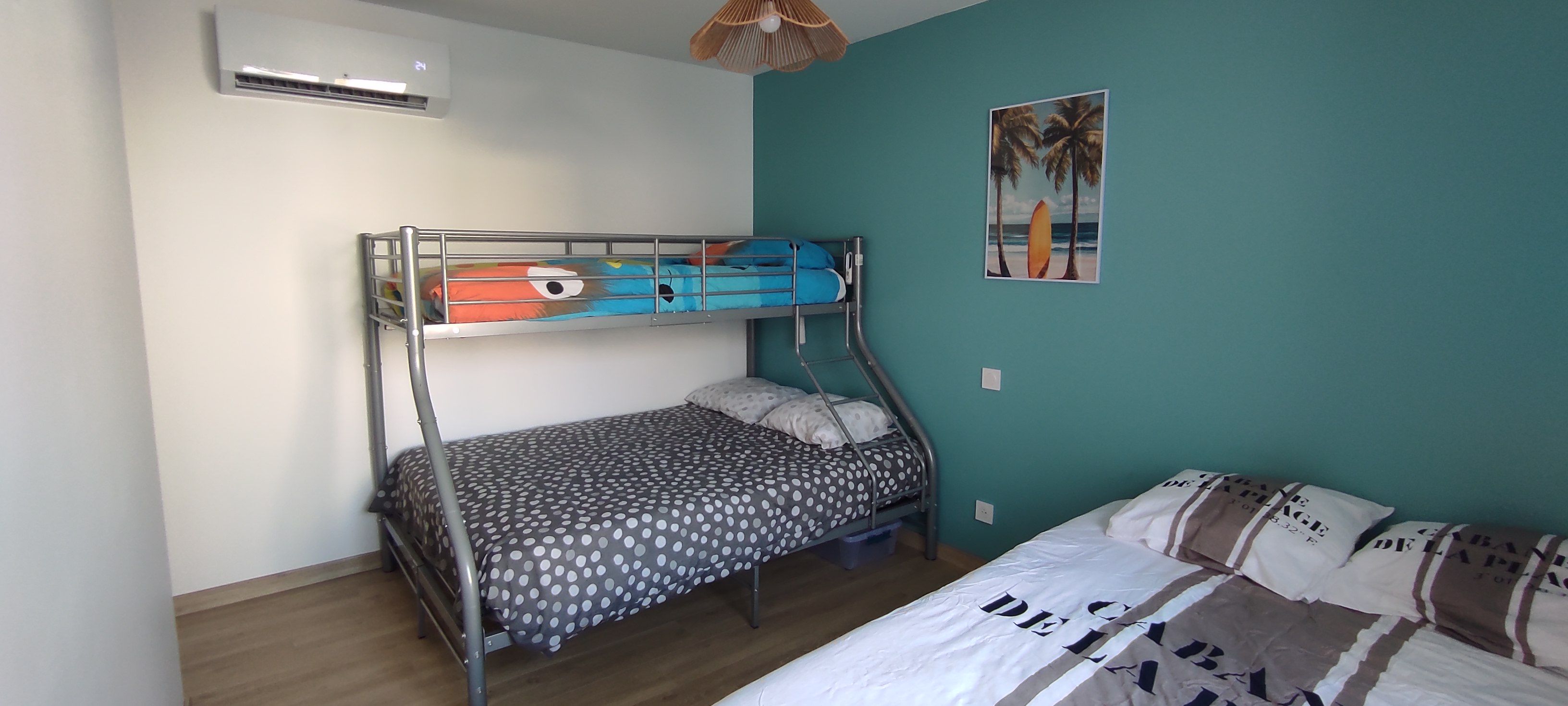 A room with a bunk bed, a single bed, a wall-mounted air conditioner, a ceiling fan, and a beach-themed painting.